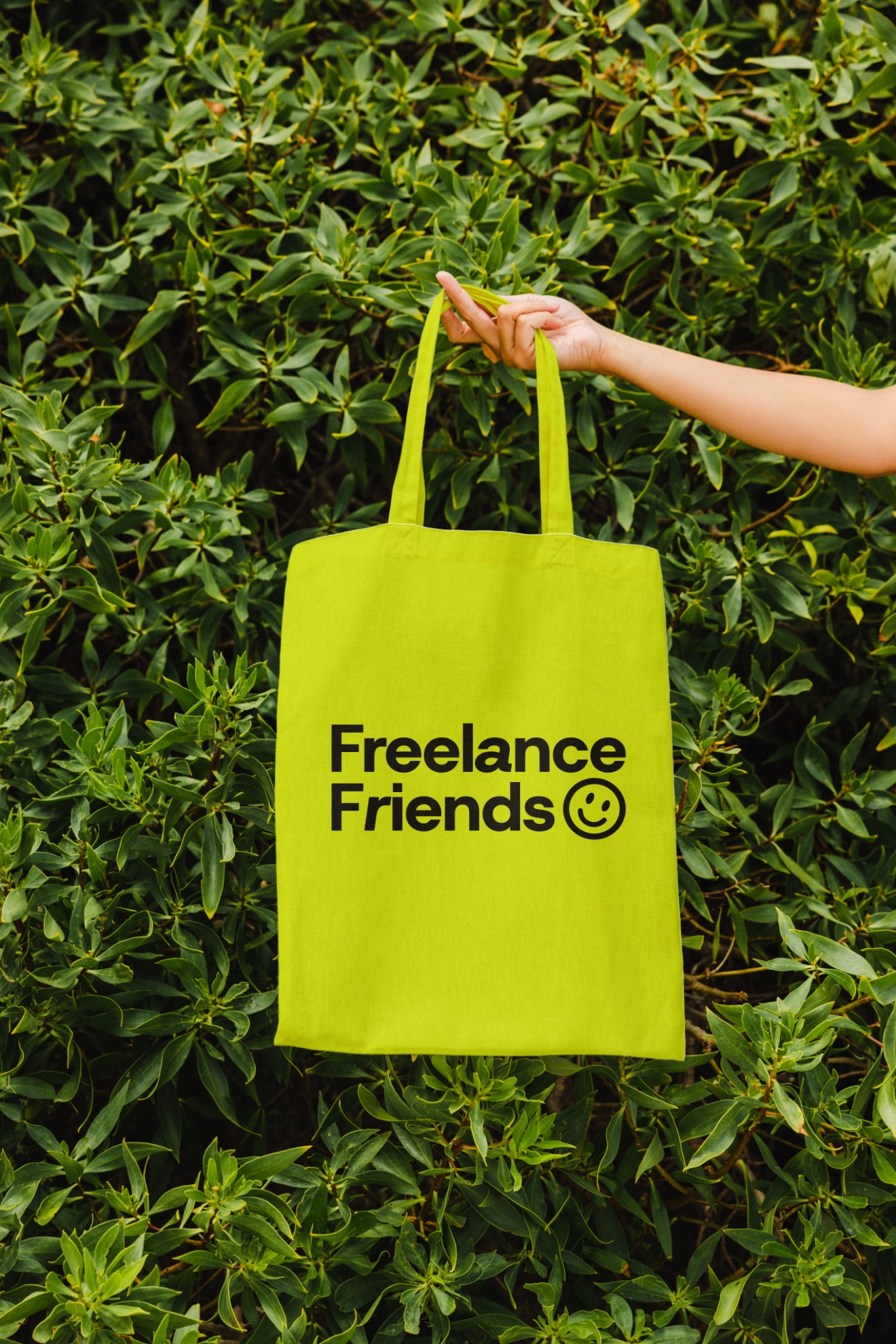 A lime Freelance Friends tote bag held against a hedge